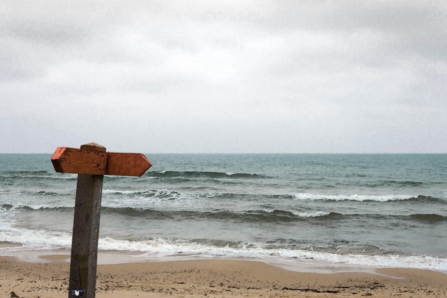 Bilden är ett fotografi av en vägskylt, där texten försvunnit. Skylten står i strandkanten, med havet som bakgrund.